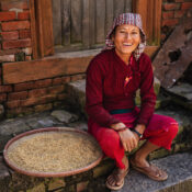 A person in a red traditional outfit and checkered headscarf smiles, sitting by a brick wall with a bamboo tray of grains. The scene conveys warmth and cultural richness.