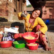 Smiling woman in traditional clothing sitting on stone steps beside a tray of harvested grains, in a rural village setting.